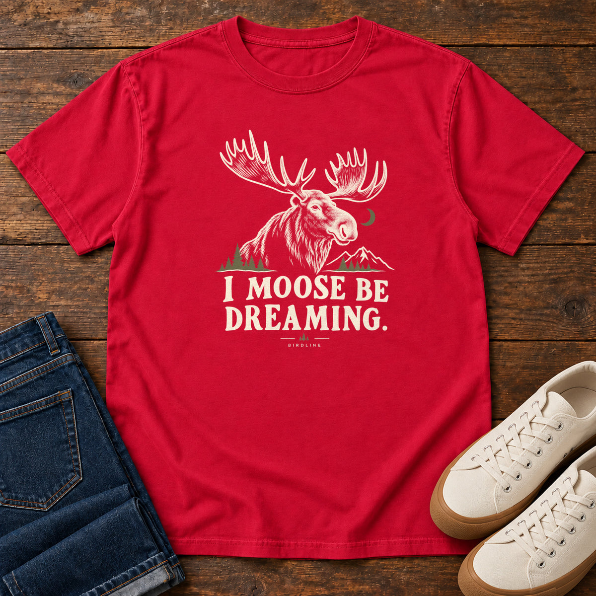 Red I Moose Be Dreaming T-Shirt with a cream moose portrait with broad antlers, mountains and a crescent moon; front flat lay on wood.