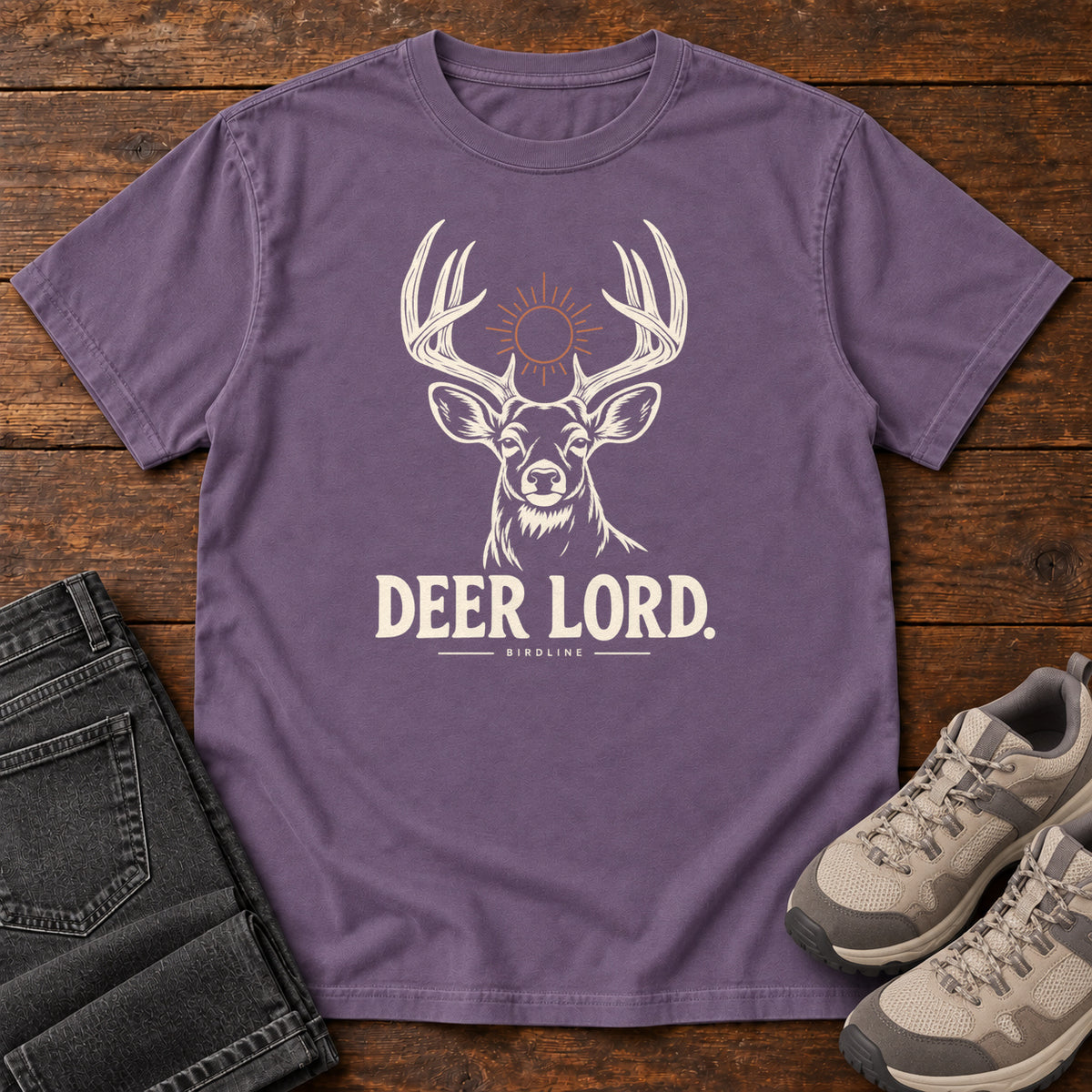 Grape Deer Lord T-Shirt with a cream antlered deer head, a sun symbol and Deer Lord lettering; front flat lay on wood.