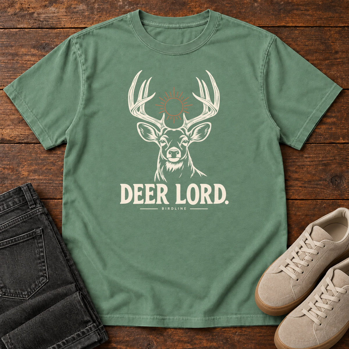 Light Green Deer Lord T-Shirt with a cream antlered deer head, a sun symbol and Deer Lord lettering; front flat lay on wood.