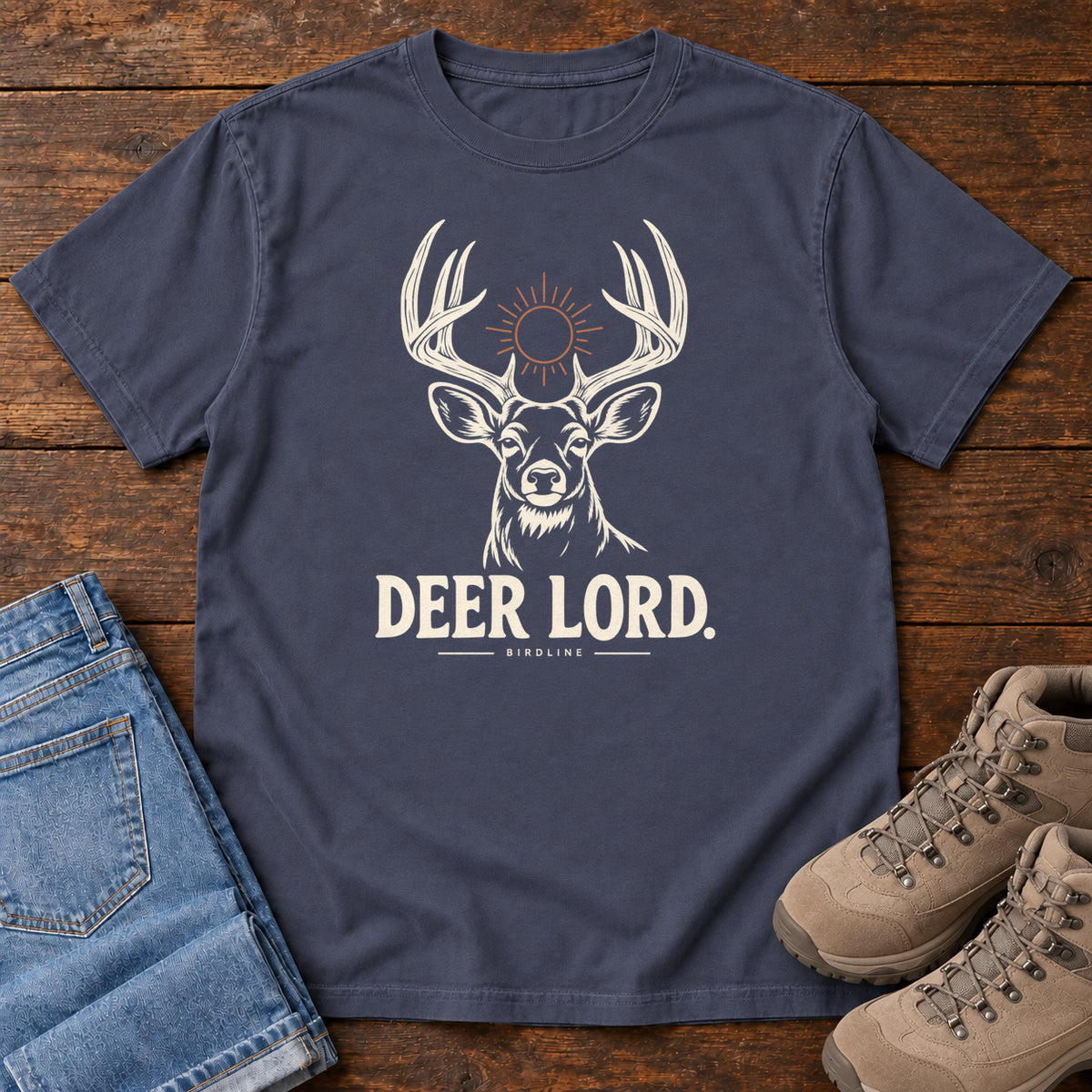 Navy Deer Lord T-Shirt with a cream antlered deer head, a sun symbol and Deer Lord lettering; front flat lay on wood.
