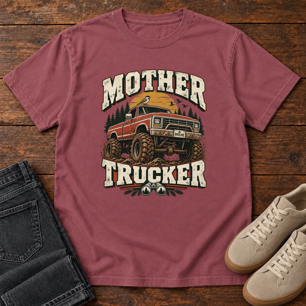 Brick Mother Trucker Red Truck T-Shirt with a lifted red pickup truck, evergreen trees and cream lettering, front flat lay on wood.