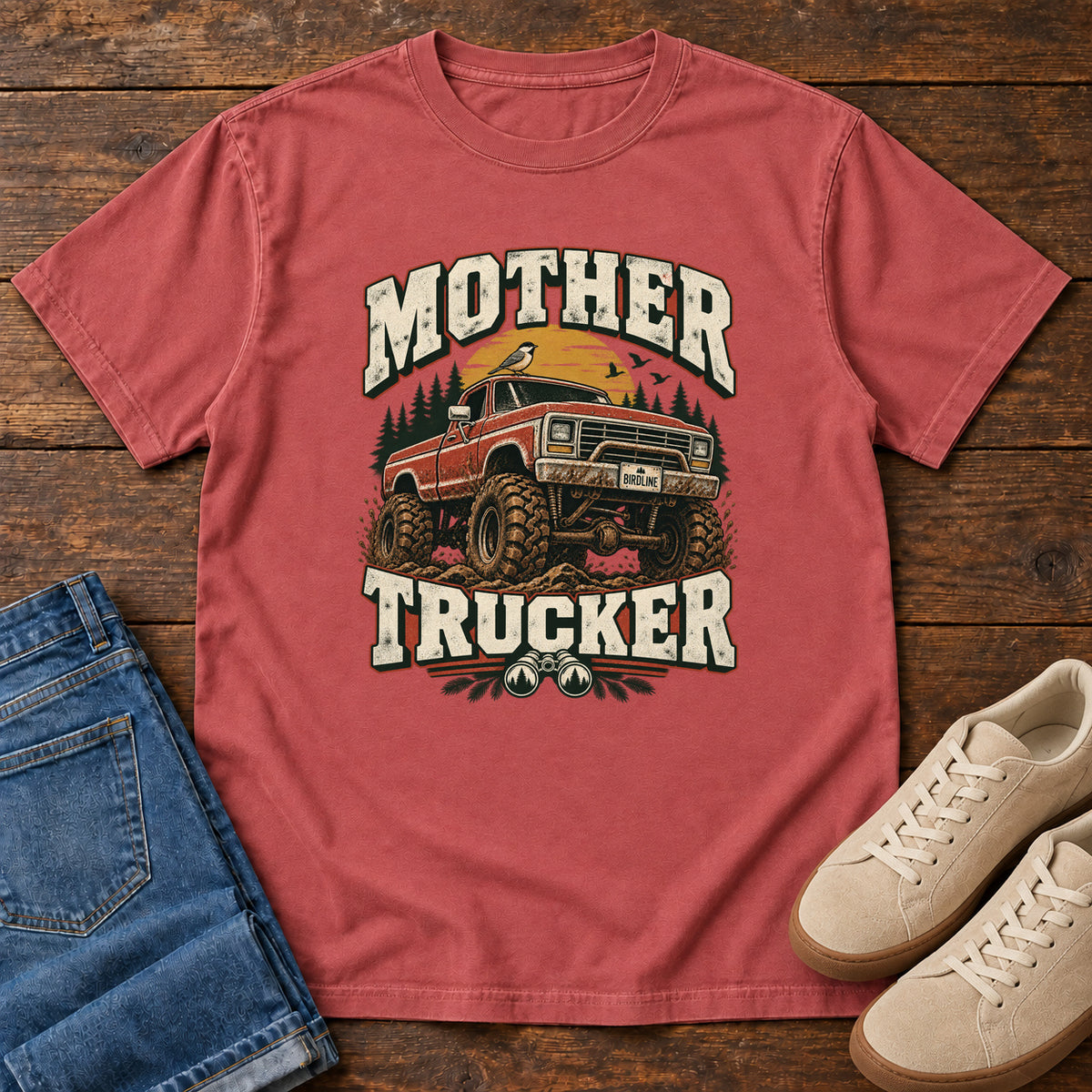 Crimson Mother Trucker Red Truck T-Shirt with a lifted red pickup truck, evergreen trees and cream lettering, front flat lay on wood.