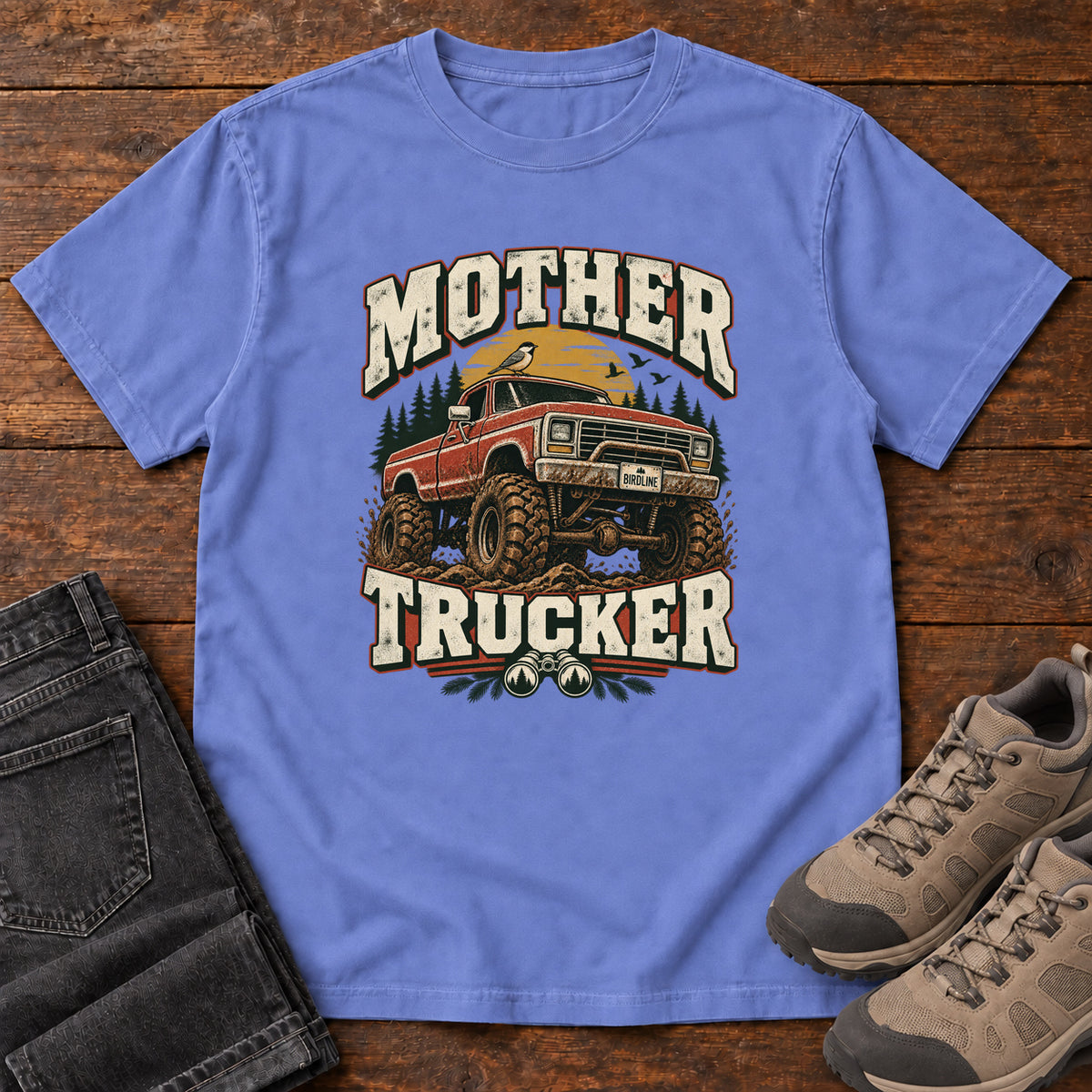 Flo Blue Mother Trucker Red Truck T-Shirt with a lifted red pickup truck, evergreen trees and cream lettering, front flat lay on wood.