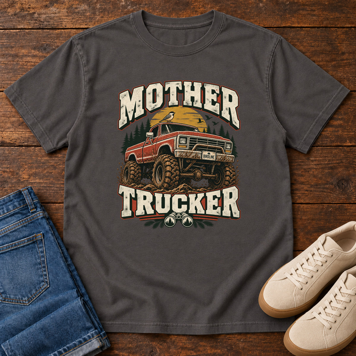 Graphite Mother Trucker Red Truck T-Shirt with a lifted red pickup truck, evergreen trees and cream lettering, front flat lay on wood.