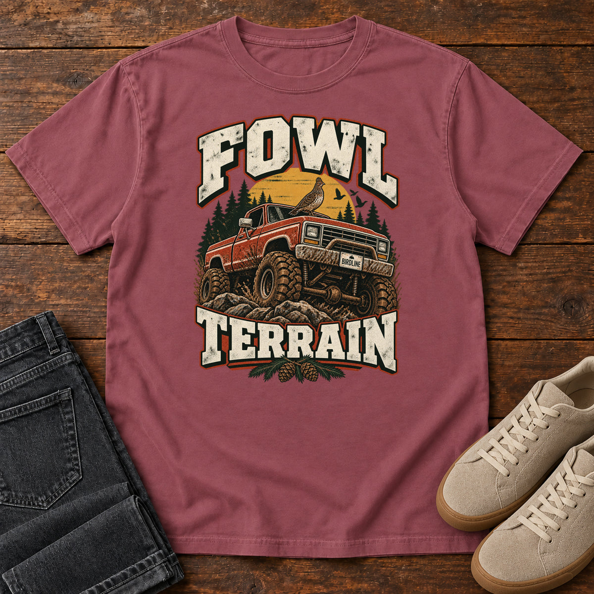 Fowl Terrain T-Shirt in Brick, front laid flat on wood, featuring an orange off-road truck with a bird on its roof, pine trees, a yellow sun and bold cream lettering.