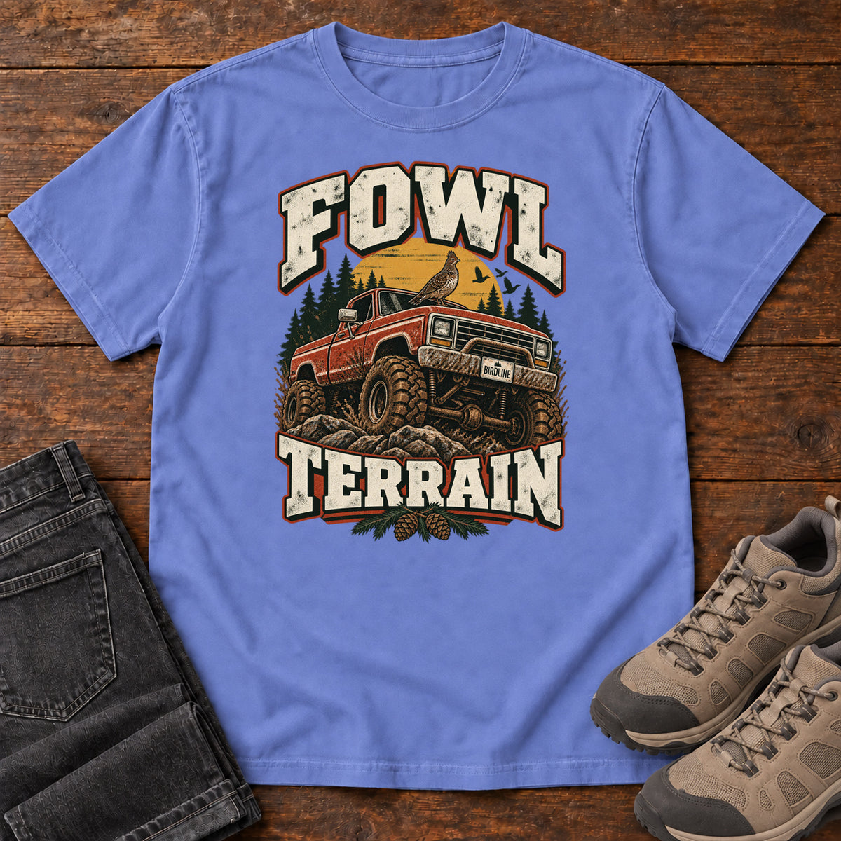 Fowl Terrain T-Shirt in Flo Blue, front laid flat on wood, featuring an orange off-road truck with a bird on its roof, pine trees, a yellow sun and bold cream lettering.