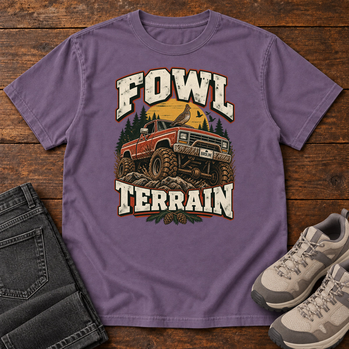 Fowl Terrain T-Shirt in Grape, front laid flat on wood, featuring an orange off-road truck with a bird on its roof, pine trees, a yellow sun and bold cream lettering.