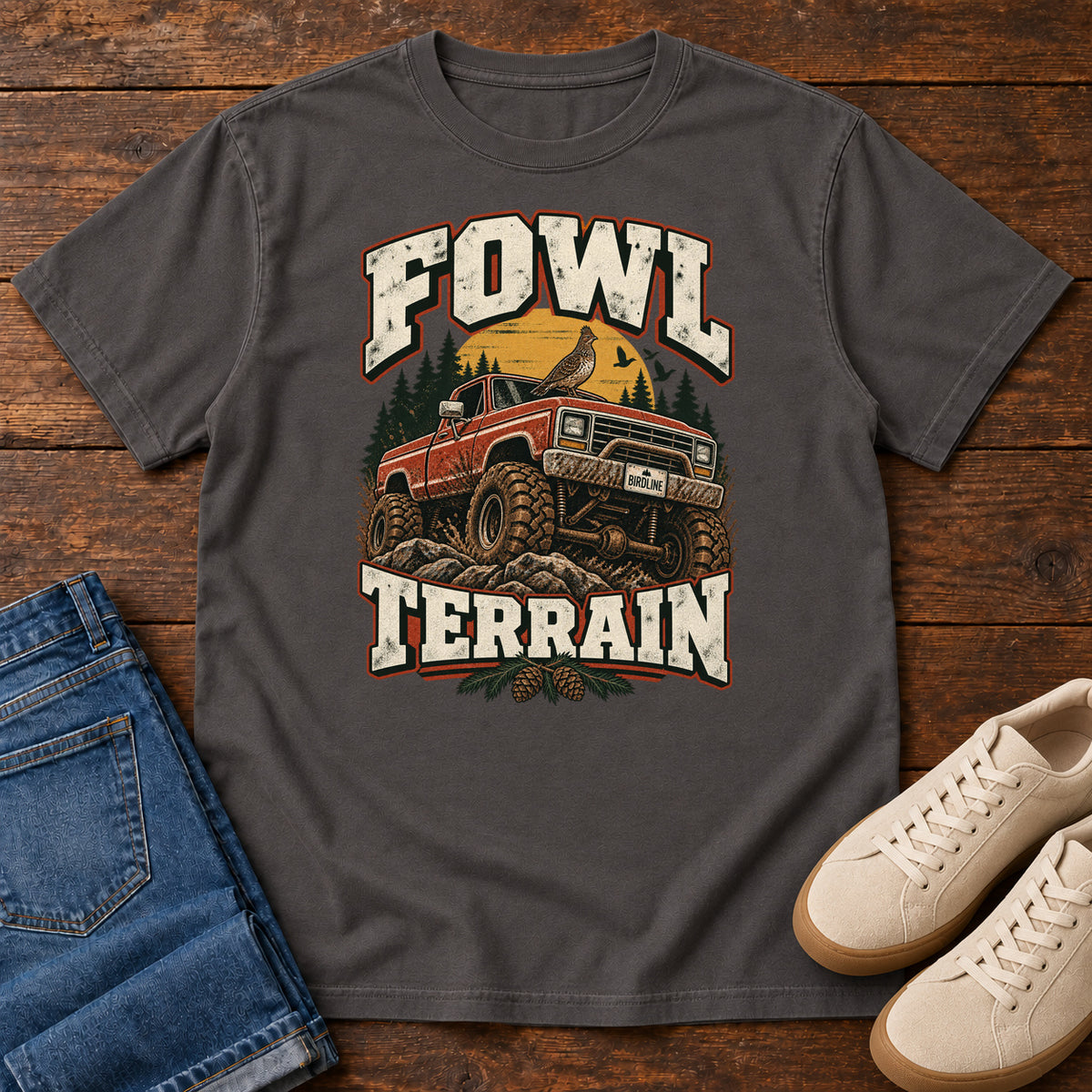 Fowl Terrain T-Shirt in Graphite, front laid flat on wood, featuring an orange off-road truck with a bird on its roof, pine trees, a yellow sun and bold cream lettering.