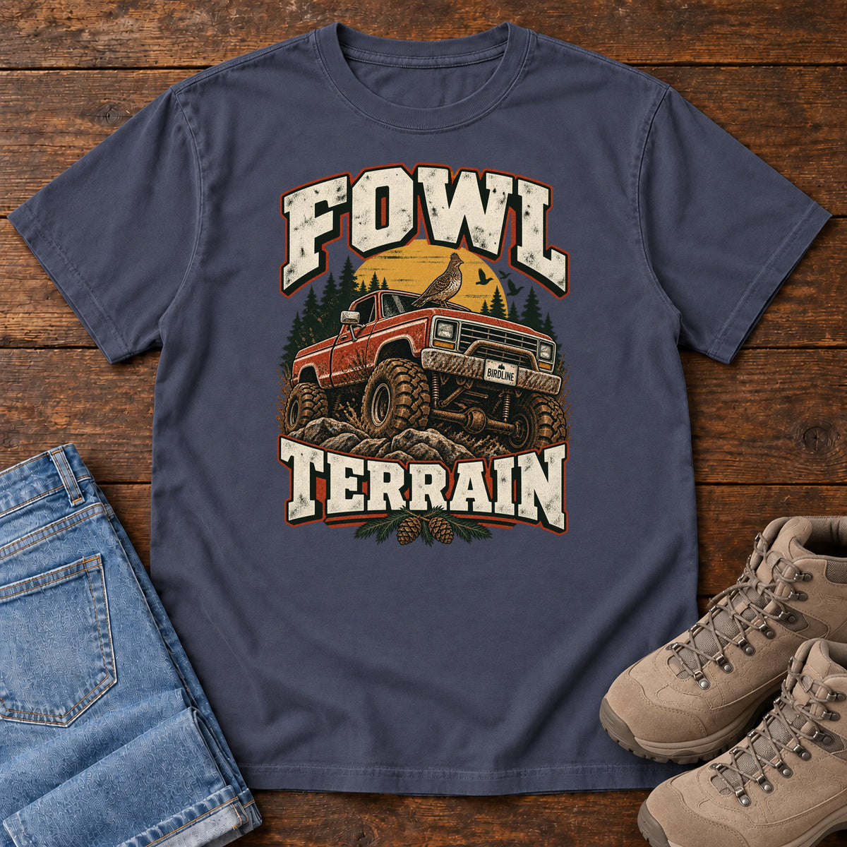 Fowl Terrain T-Shirt in Navy, front laid flat on wood, featuring an orange off-road truck with a bird on its roof, pine trees, a yellow sun and bold cream lettering.