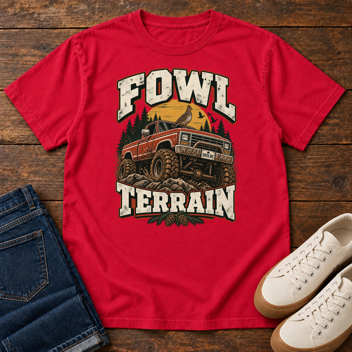 Fowl Terrain T-Shirt in Red, front laid flat on wood, featuring an orange off-road truck with a bird on its roof, pine trees, a yellow sun and bold cream lettering.