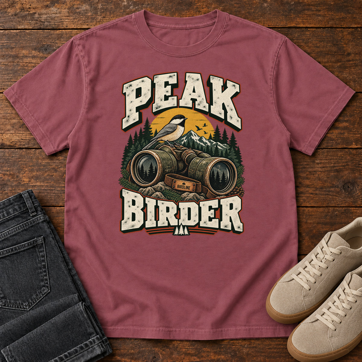 Brick Peak Birder T-Shirt with a small black-capped bird on binoculars, with mountains and distressed cream lettering, front flat lay on wood.