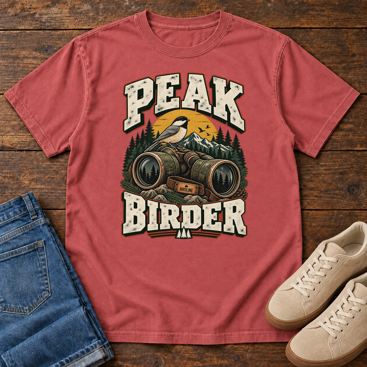 Crimson Peak Birder T-Shirt with a small black-capped bird on binoculars, with mountains and distressed cream lettering, front flat lay on wood.