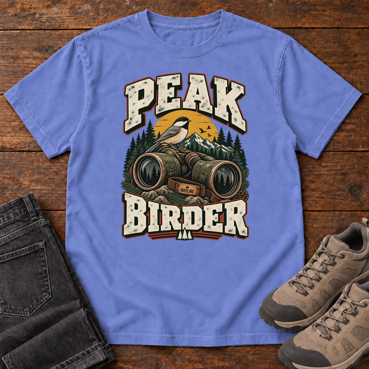 Flo Blue Peak Birder T-Shirt with a small black-capped bird on binoculars, with mountains and distressed cream lettering, front flat lay on wood.