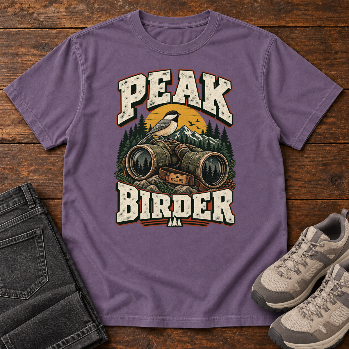 Grape Peak Birder T-Shirt with a small black-capped bird on binoculars, with mountains and distressed cream lettering, front flat lay on wood.