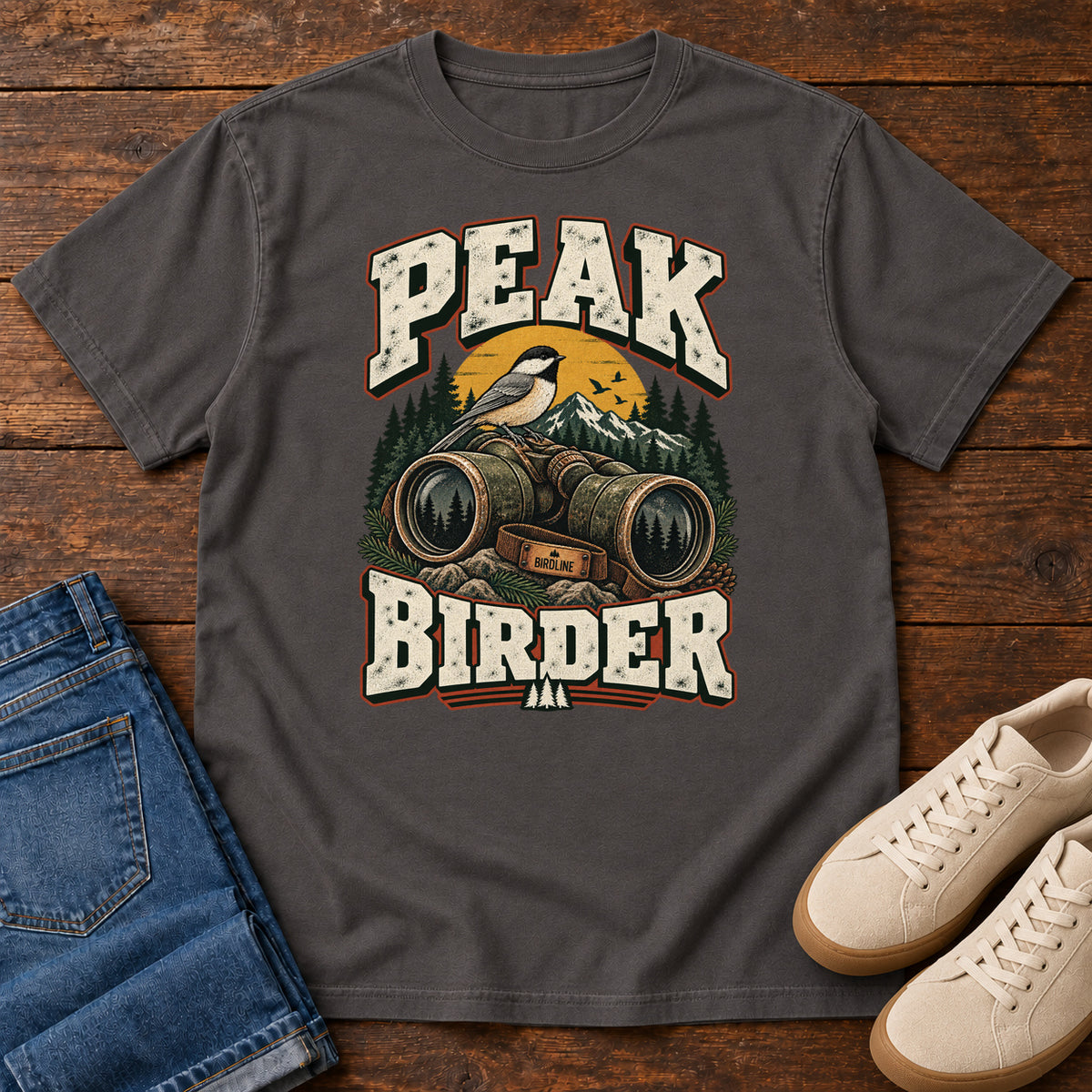 Graphite Peak Birder T-Shirt with a small black-capped bird on binoculars, with mountains and distressed cream lettering, front flat lay on wood.