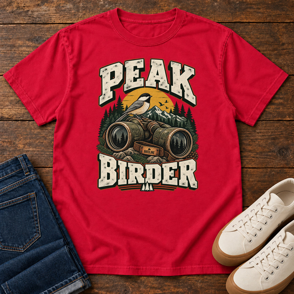 Red Peak Birder T-Shirt with a small black-capped bird on binoculars, with mountains and distressed cream lettering, front flat lay on wood.