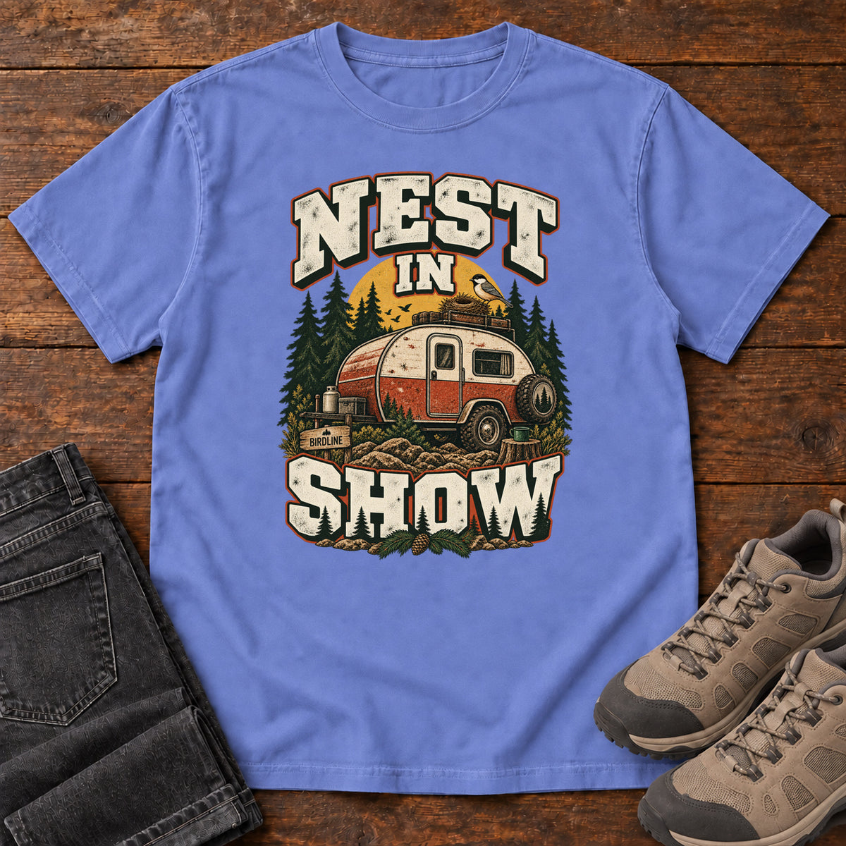 Flo Blue Nest in Show T-Shirt with a red-and-cream camper beneath pines, with a small bird and nest on its roof; front flat lay on wood.