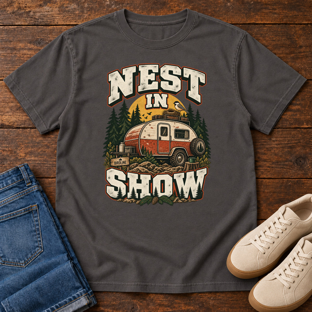 Graphite Nest in Show T-Shirt with a red-and-cream camper beneath pines, with a small bird and nest on its roof; front flat lay on wood.