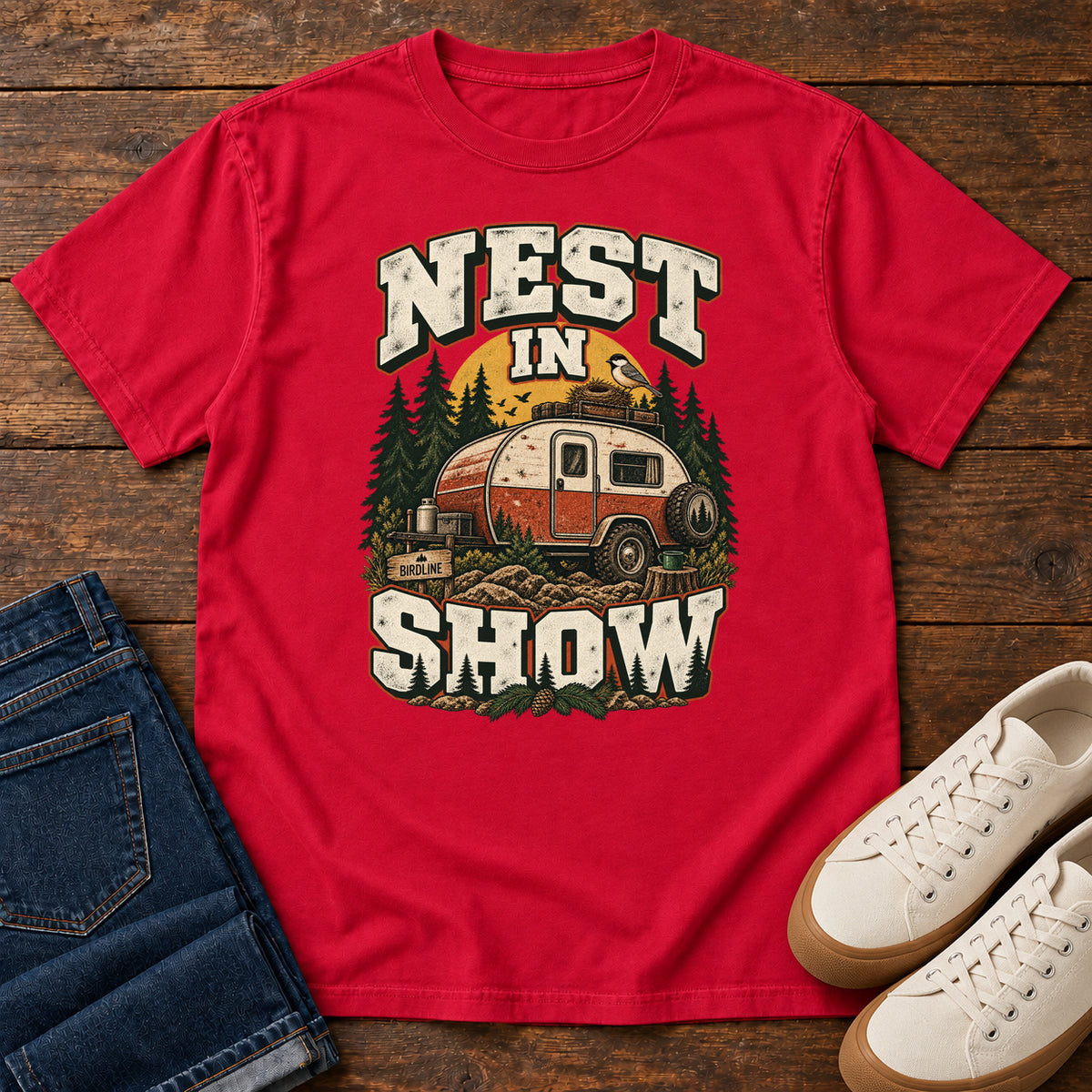 Red Nest in Show T-Shirt with a red-and-cream camper beneath pines, with a small bird and nest on its roof; front flat lay on wood.