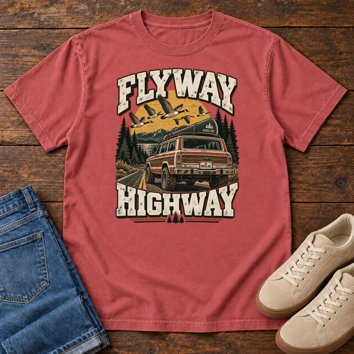 Crimson Flyway Highway T-Shirt with a vintage pickup on a forest road with flying birds overhead; front flat lay on wood.