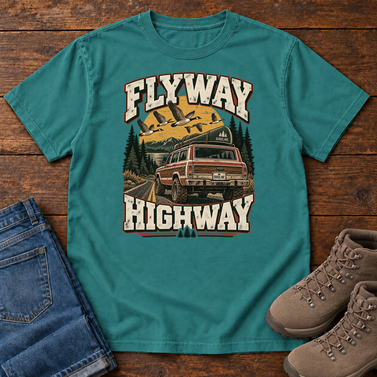 Emerald Flyway Highway T-Shirt with a vintage pickup on a forest road with flying birds overhead; front flat lay on wood.
