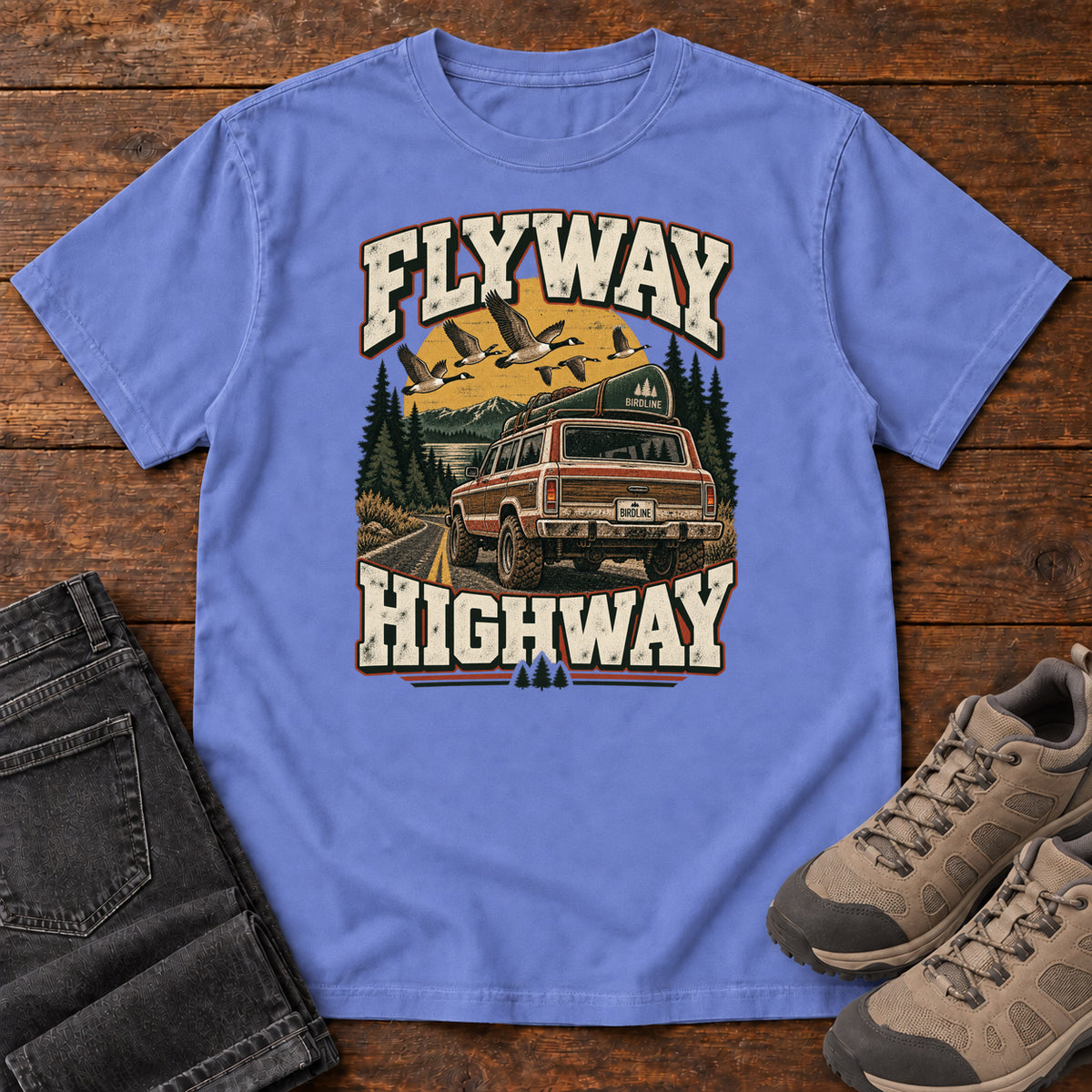 Flo Blue Flyway Highway T-Shirt with a vintage pickup on a forest road with flying birds overhead; front flat lay on wood.