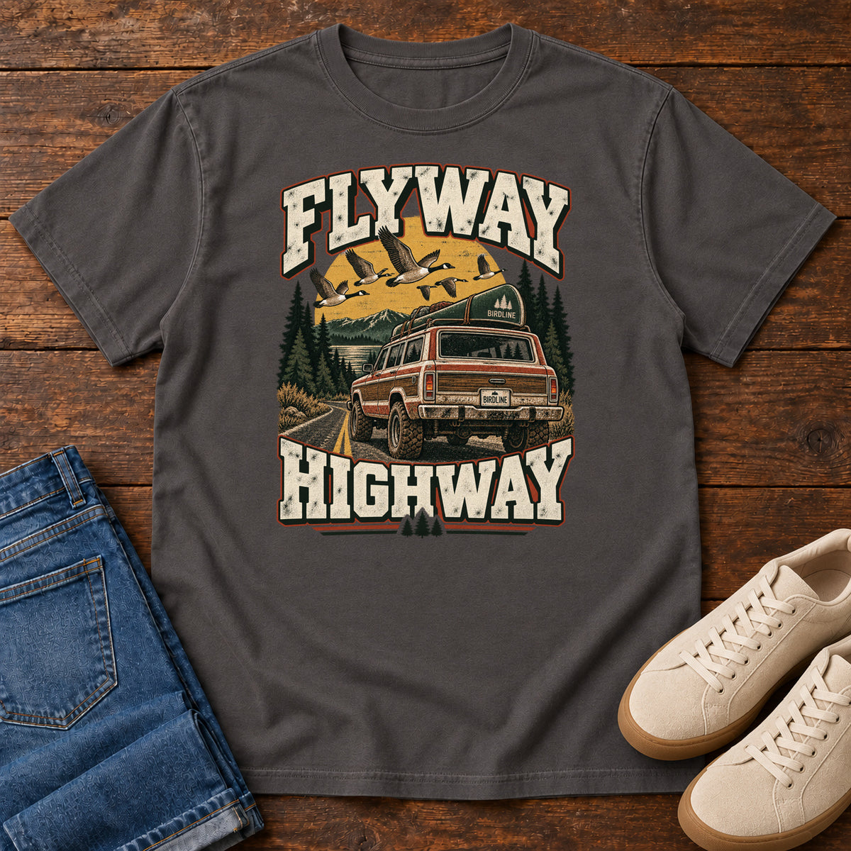 Graphite Flyway Highway T-Shirt with a vintage pickup on a forest road with flying birds overhead; front flat lay on wood.