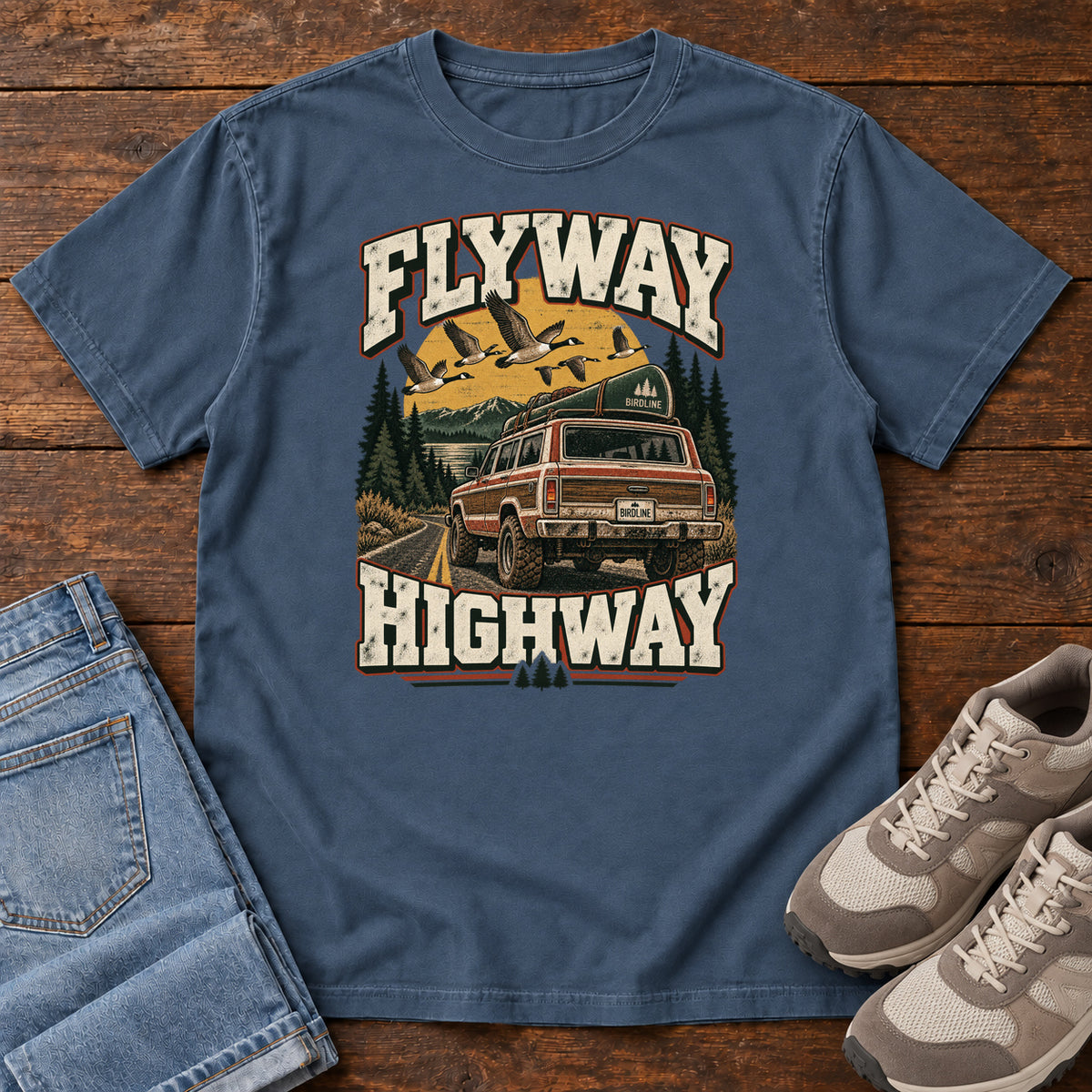 Midnight Flyway Highway T-Shirt with a vintage pickup on a forest road with flying birds overhead; front flat lay on wood.