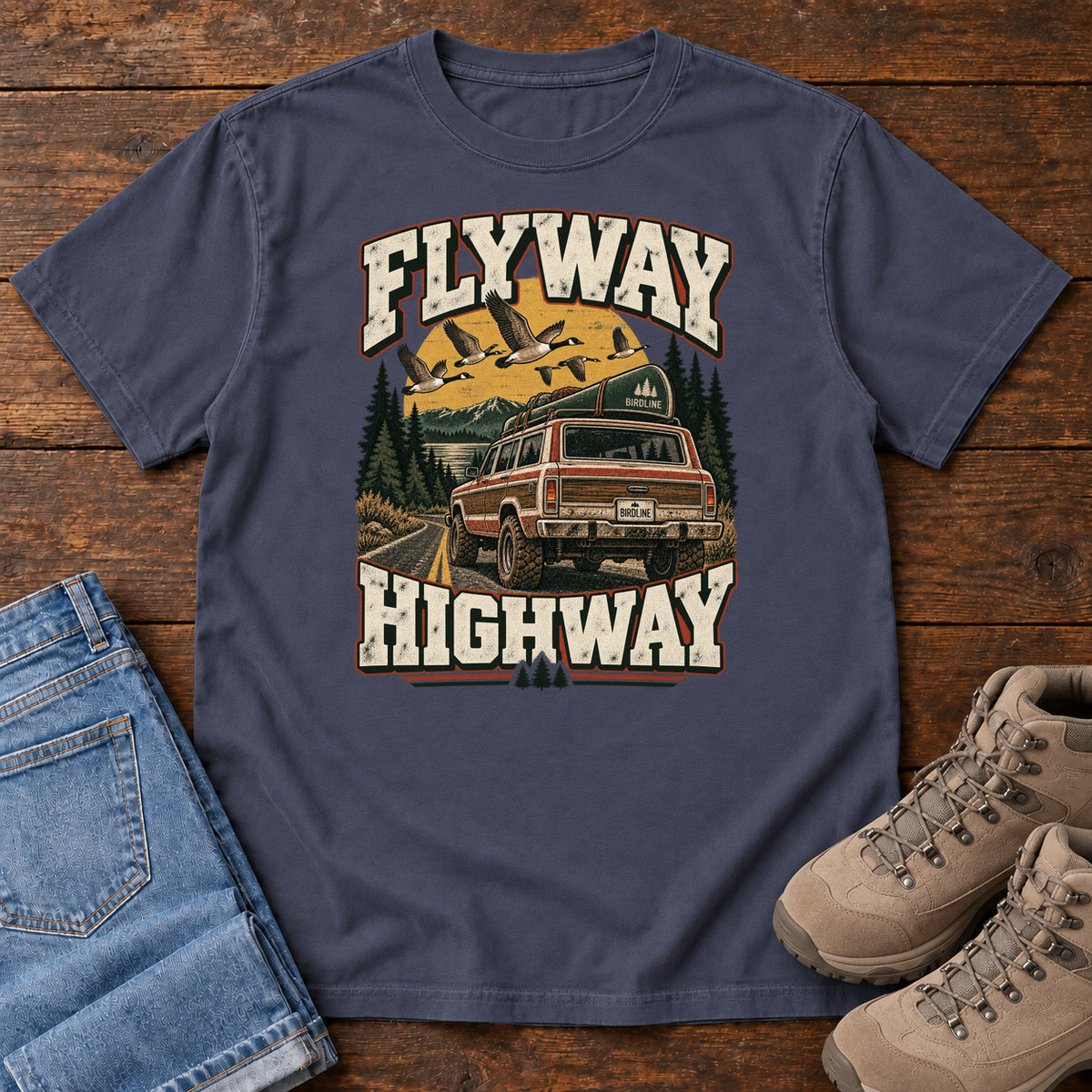 Navy Flyway Highway T-Shirt with a vintage pickup on a forest road with flying birds overhead; front flat lay on wood.