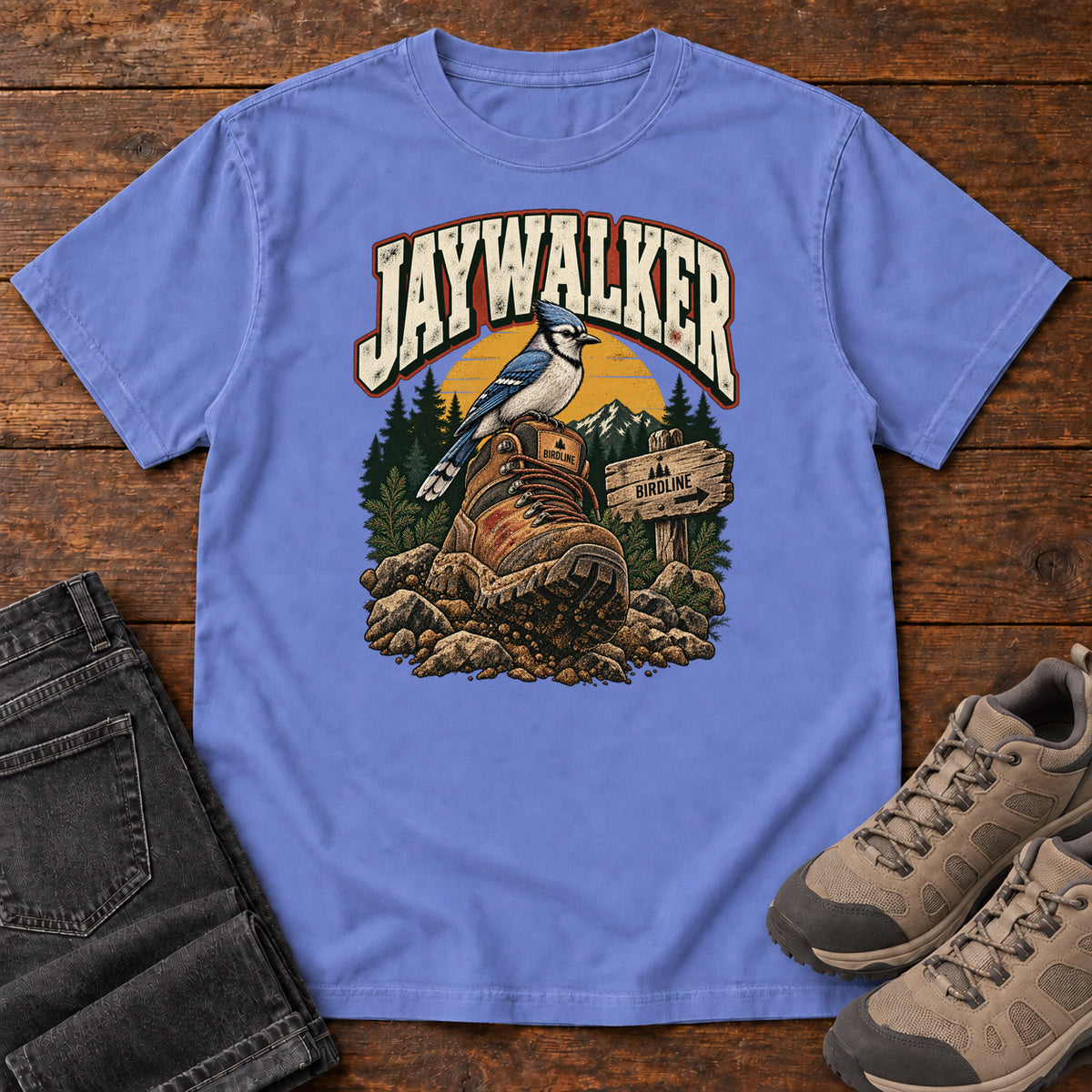 Flo Blue Jaywalker T-Shirt with a blue-and-white bird on a hiking boot beside a trail sign, front flat lay on wood.