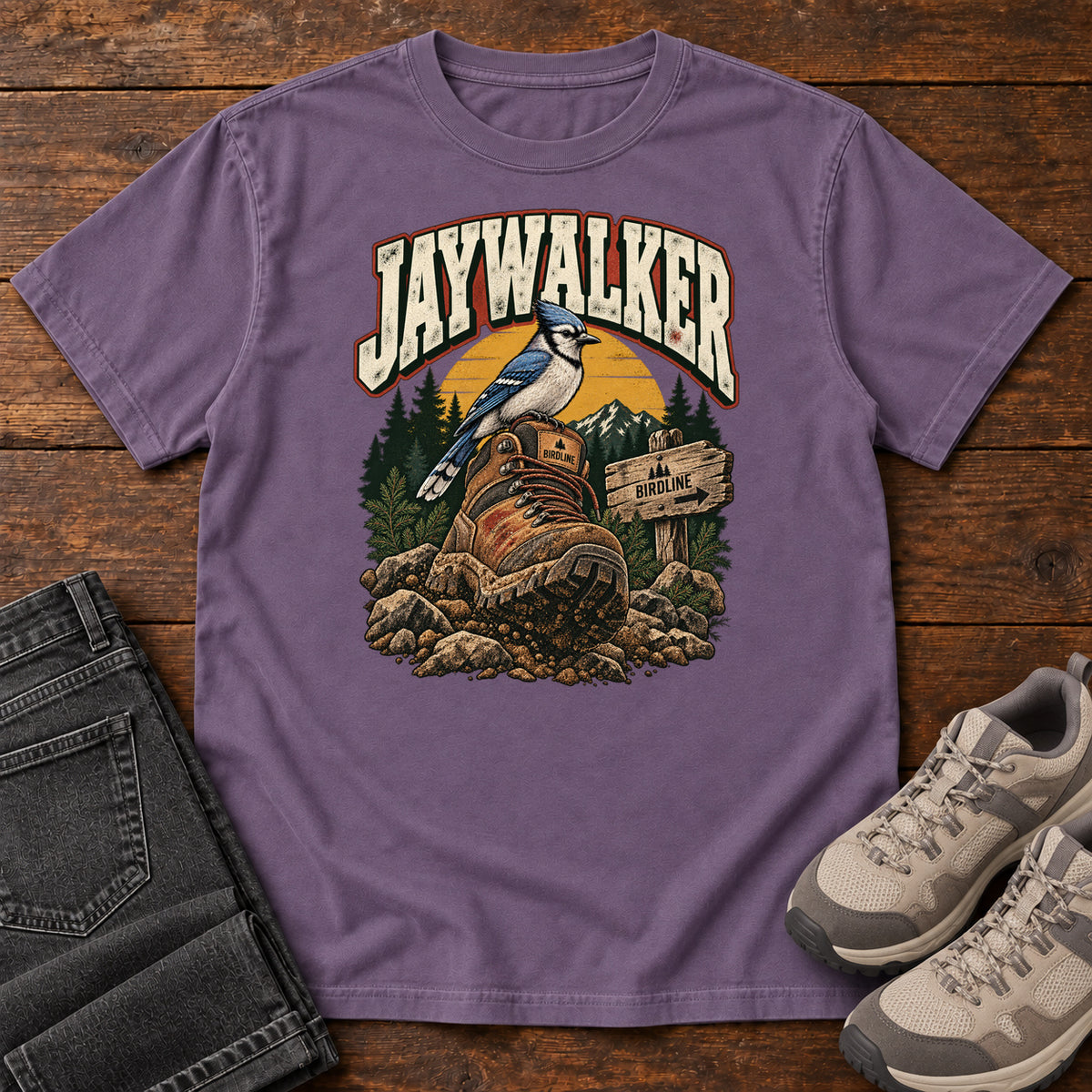 Grape Jaywalker T-Shirt with a blue-and-white bird on a hiking boot beside a trail sign, front flat lay on wood.