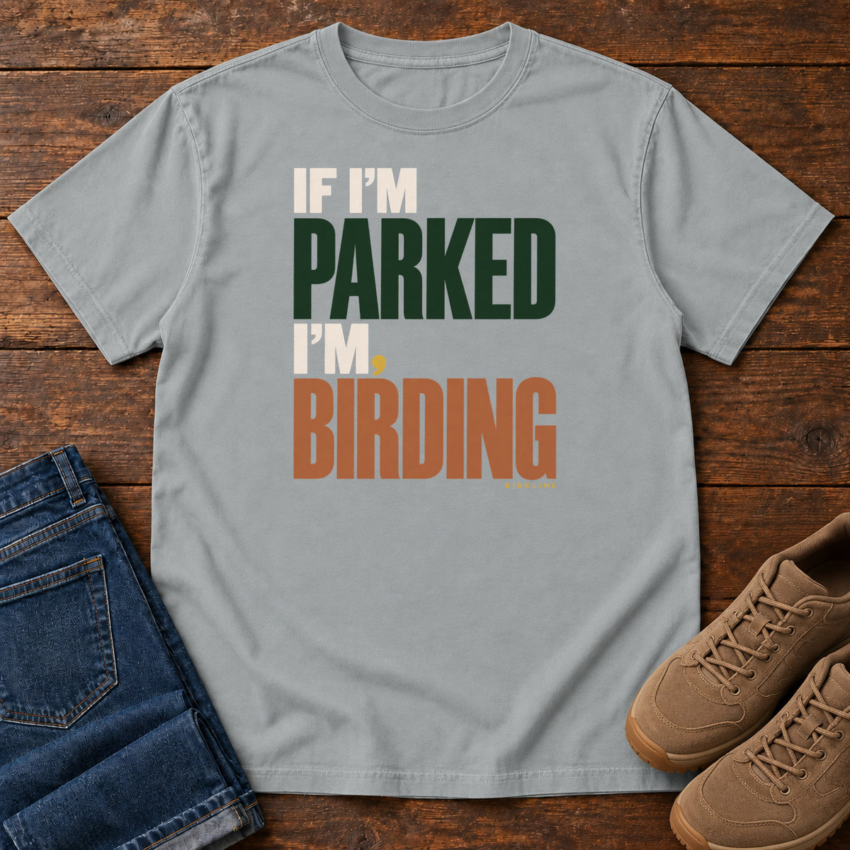 If I'm Parked I'm Birding T-Shirt in Granite, front laid flat on wood, featuring stacked cream, dark green and rust slogan lettering with a small yellow dot.