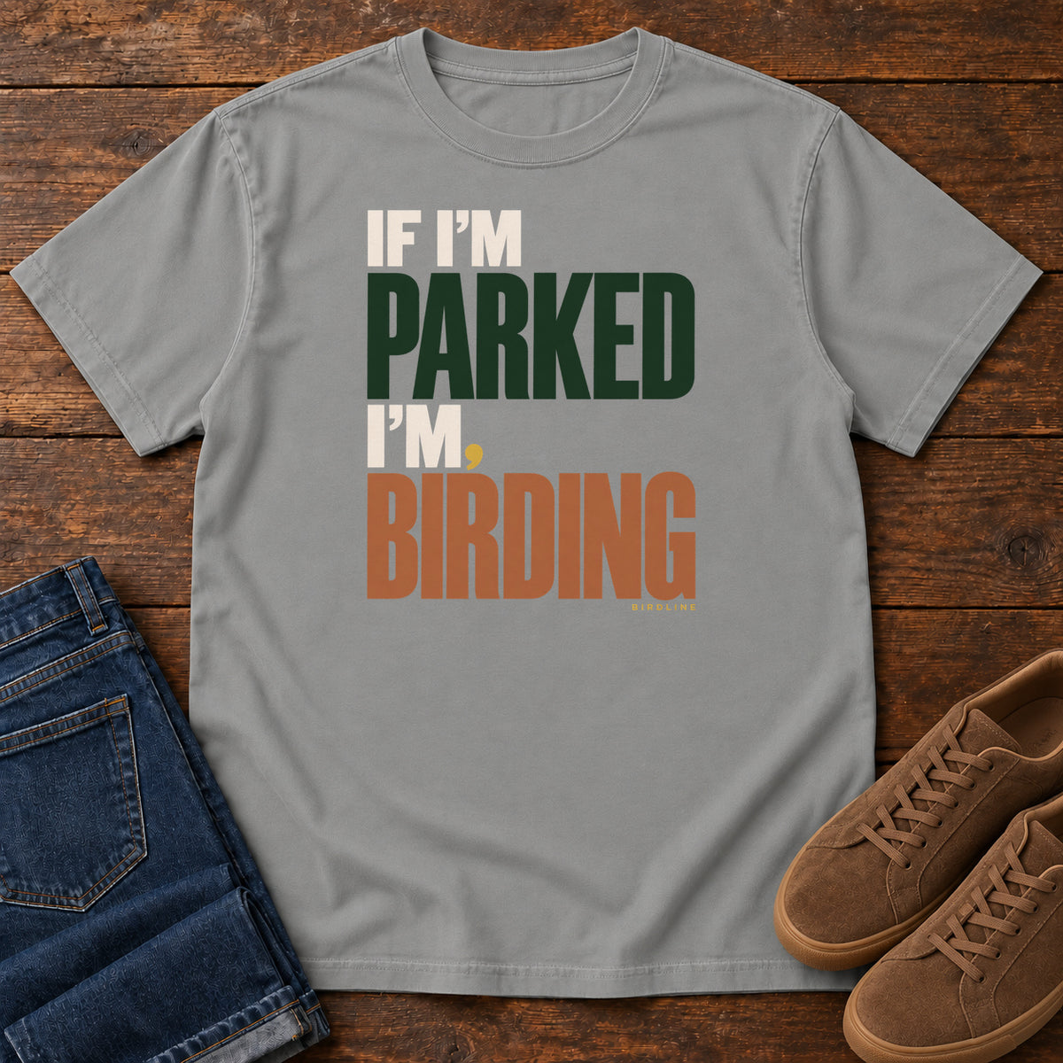If I'm Parked I'm Birding T-Shirt in Grey, front laid flat on wood, featuring stacked cream, dark green and rust slogan lettering with a small yellow dot.