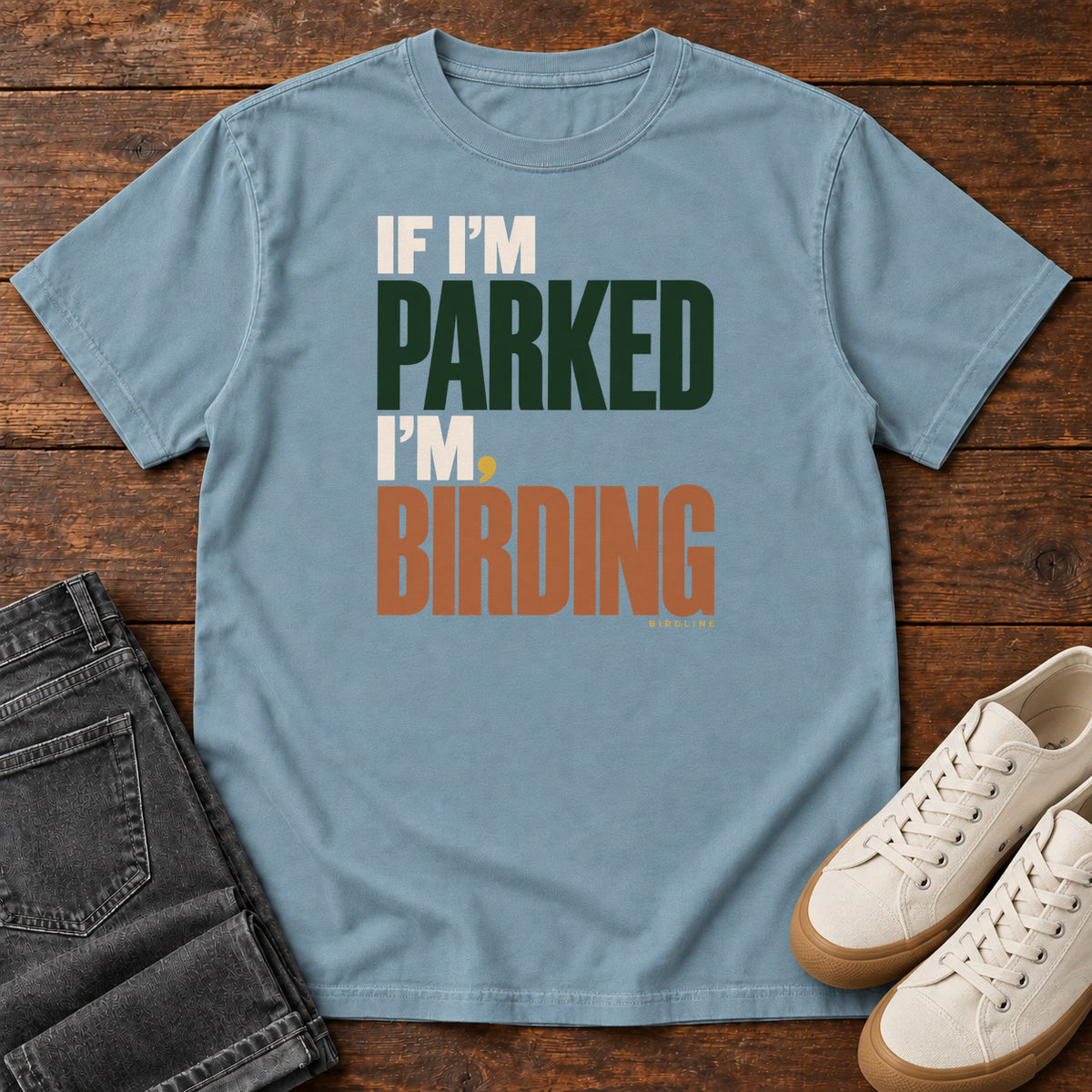 If I'm Parked I'm Birding T-Shirt in Ice Blue, front laid flat on wood, featuring stacked cream, dark green and rust slogan lettering with a small yellow dot.