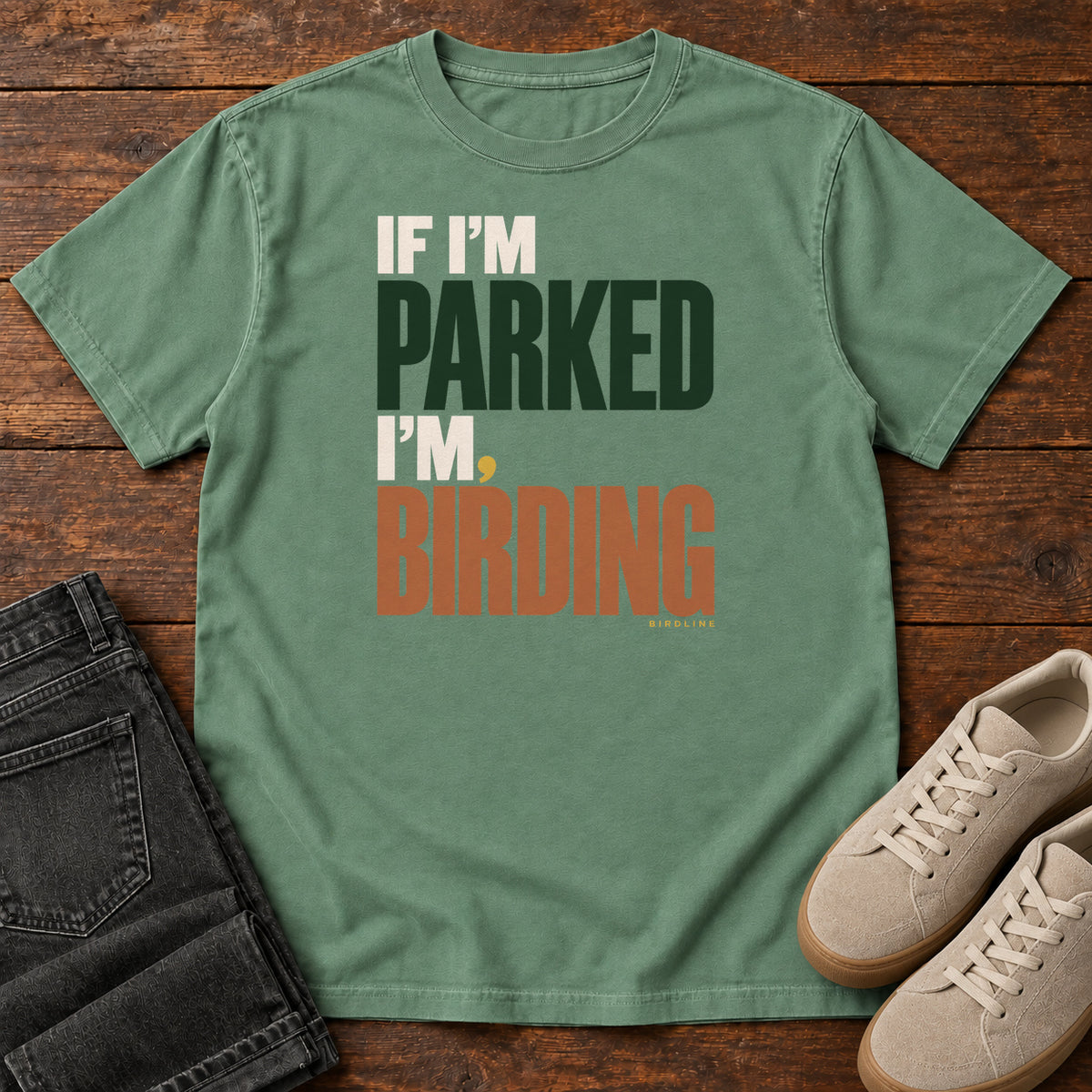 If I'm Parked I'm Birding T-Shirt in Light Green, front laid flat on wood, featuring stacked cream, dark green and rust slogan lettering with a small yellow dot.