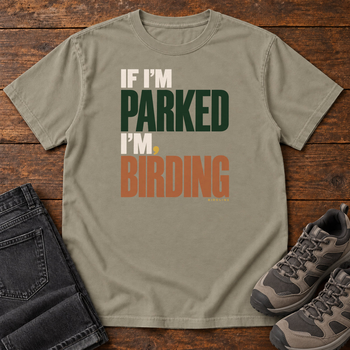 If I'm Parked I'm Birding T-Shirt in Sandstone, front laid flat on wood, featuring stacked cream, dark green and rust slogan lettering with a small yellow dot.