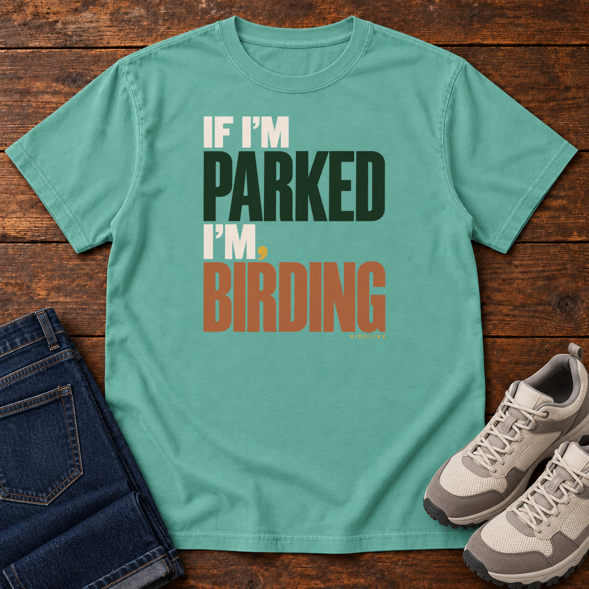 If I'm Parked I'm Birding T-Shirt in Seafoam, front laid flat on wood, featuring stacked cream, dark green and rust slogan lettering with a small yellow dot.