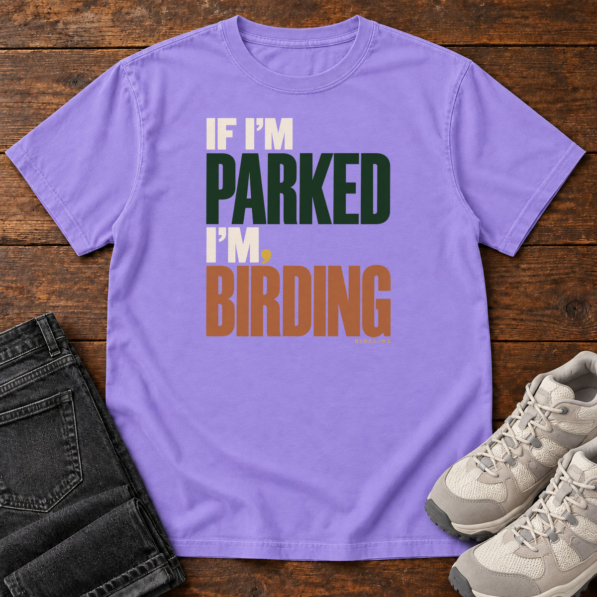 If I'm Parked I'm Birding T-Shirt in Violet, front laid flat on wood, featuring stacked cream, dark green and rust slogan lettering with a small yellow dot.