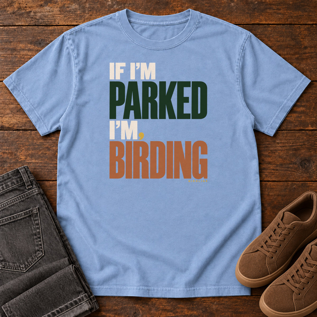 If I'm Parked I'm Birding T-Shirt in Washed Denim, front laid flat on wood, featuring stacked cream, dark green and rust slogan lettering with a small yellow dot.