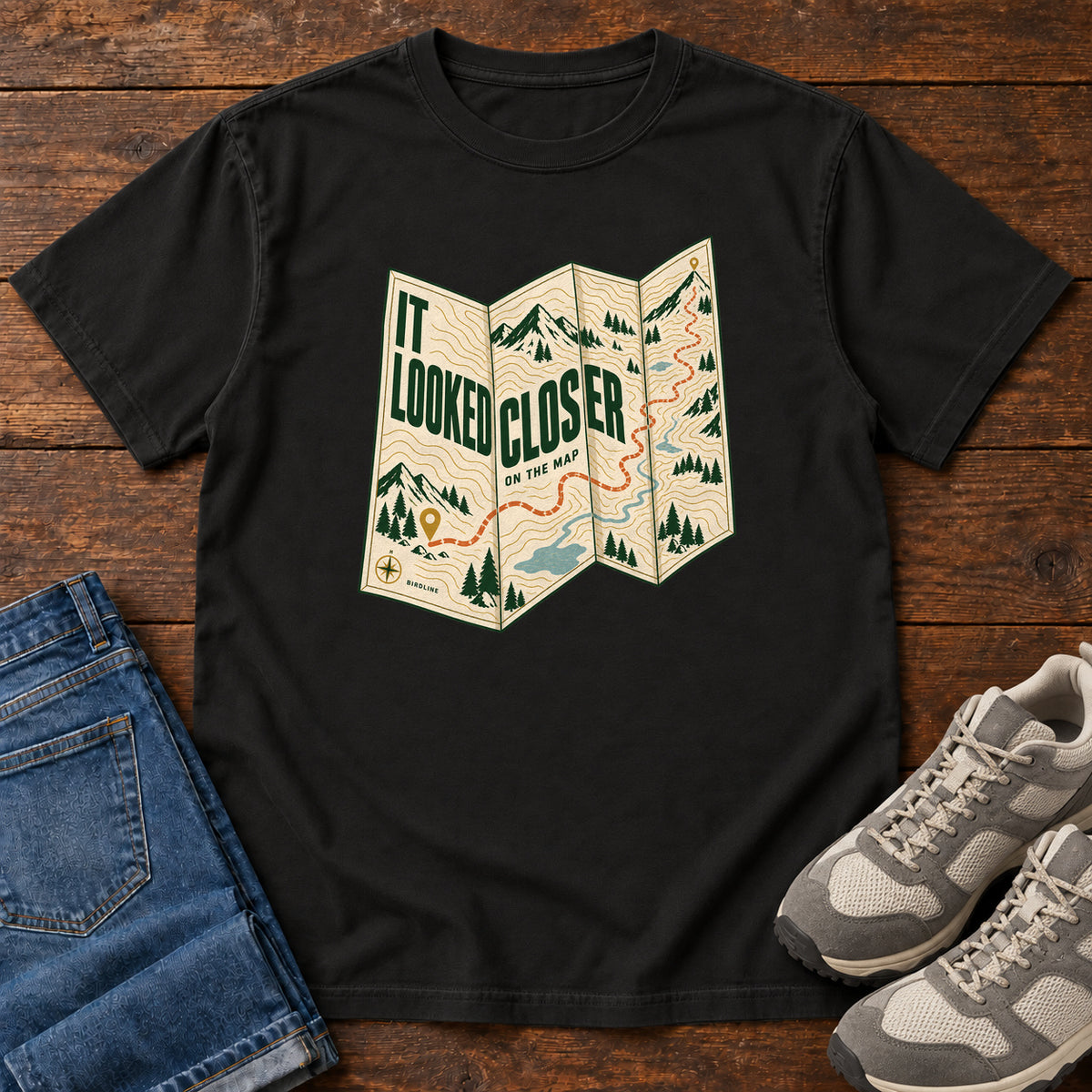 Black It Looked Closer on the Map T-Shirt with a folded cream map graphic, green lettering and an orange route, front flat lay on wood.