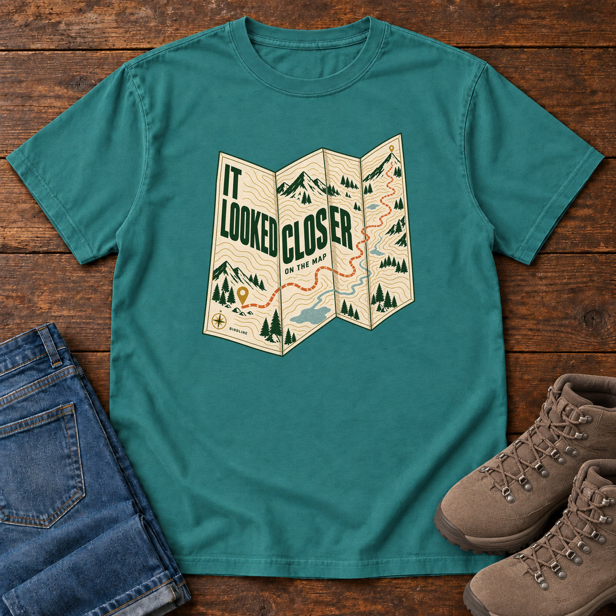 Emerald It Looked Closer on the Map T-Shirt with a folded cream map graphic, green lettering and an orange route, front flat lay on wood.