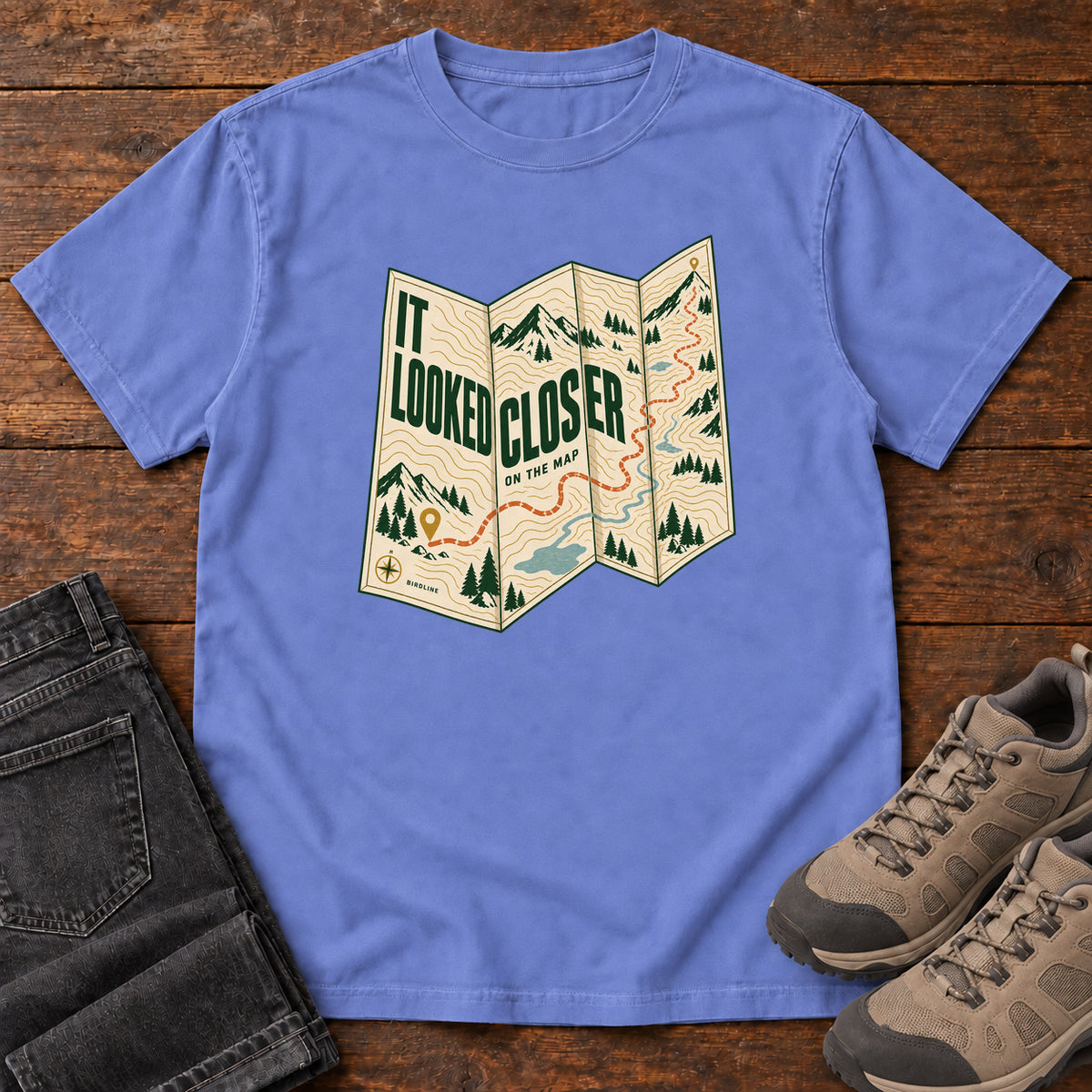 Flo Blue It Looked Closer on the Map T-Shirt with a folded cream map graphic, green lettering and an orange route, front flat lay on wood.