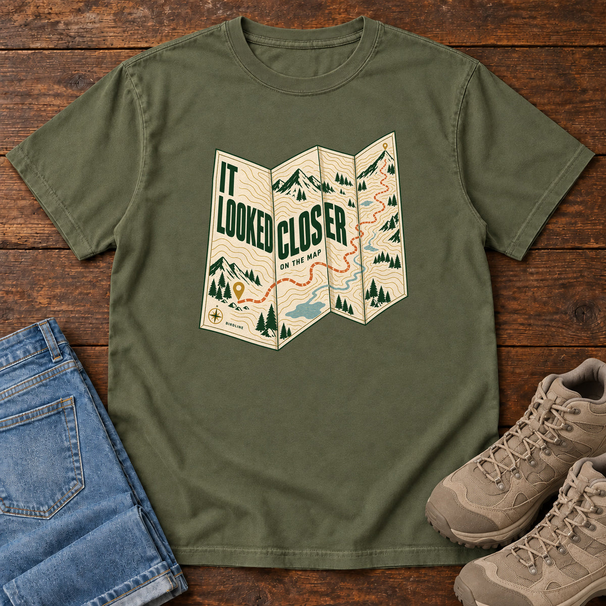 Hemp It Looked Closer on the Map T-Shirt with a folded cream map graphic, green lettering and an orange route, front flat lay on wood.