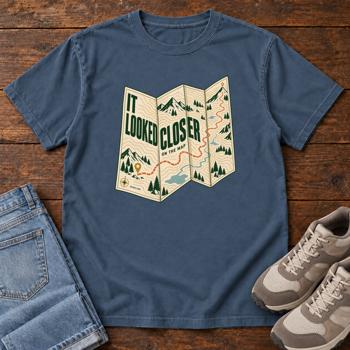 Midnight It Looked Closer on the Map T-Shirt with a folded cream map graphic, green lettering and an orange route, front flat lay on wood.