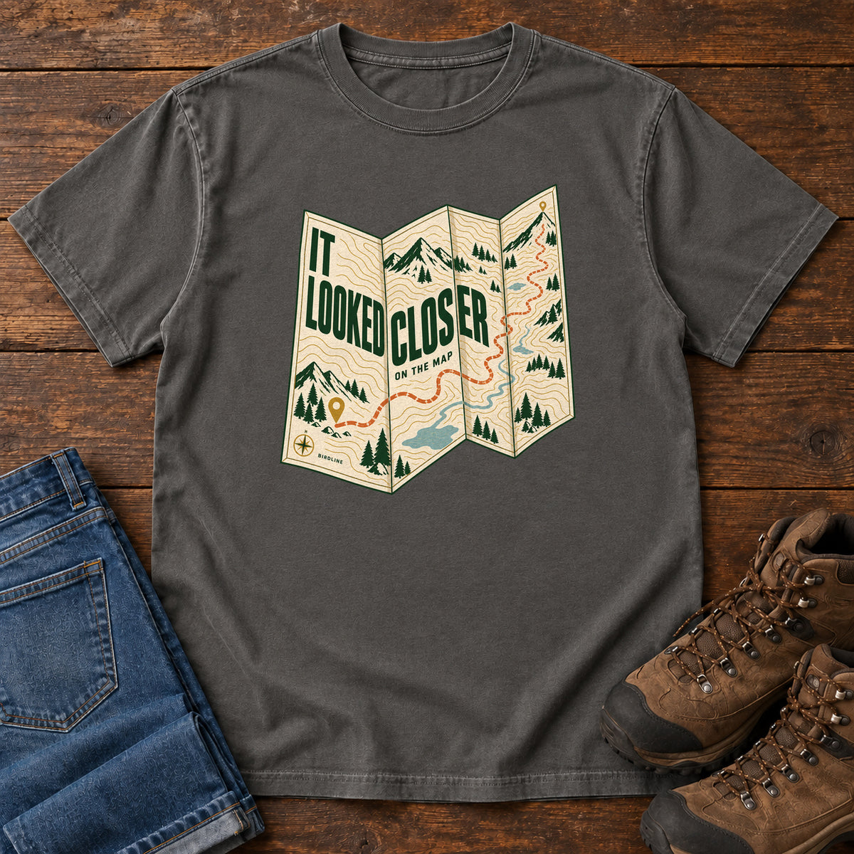 Pepper It Looked Closer on the Map T-Shirt with a folded cream map graphic, green lettering and an orange route, front flat lay on wood.