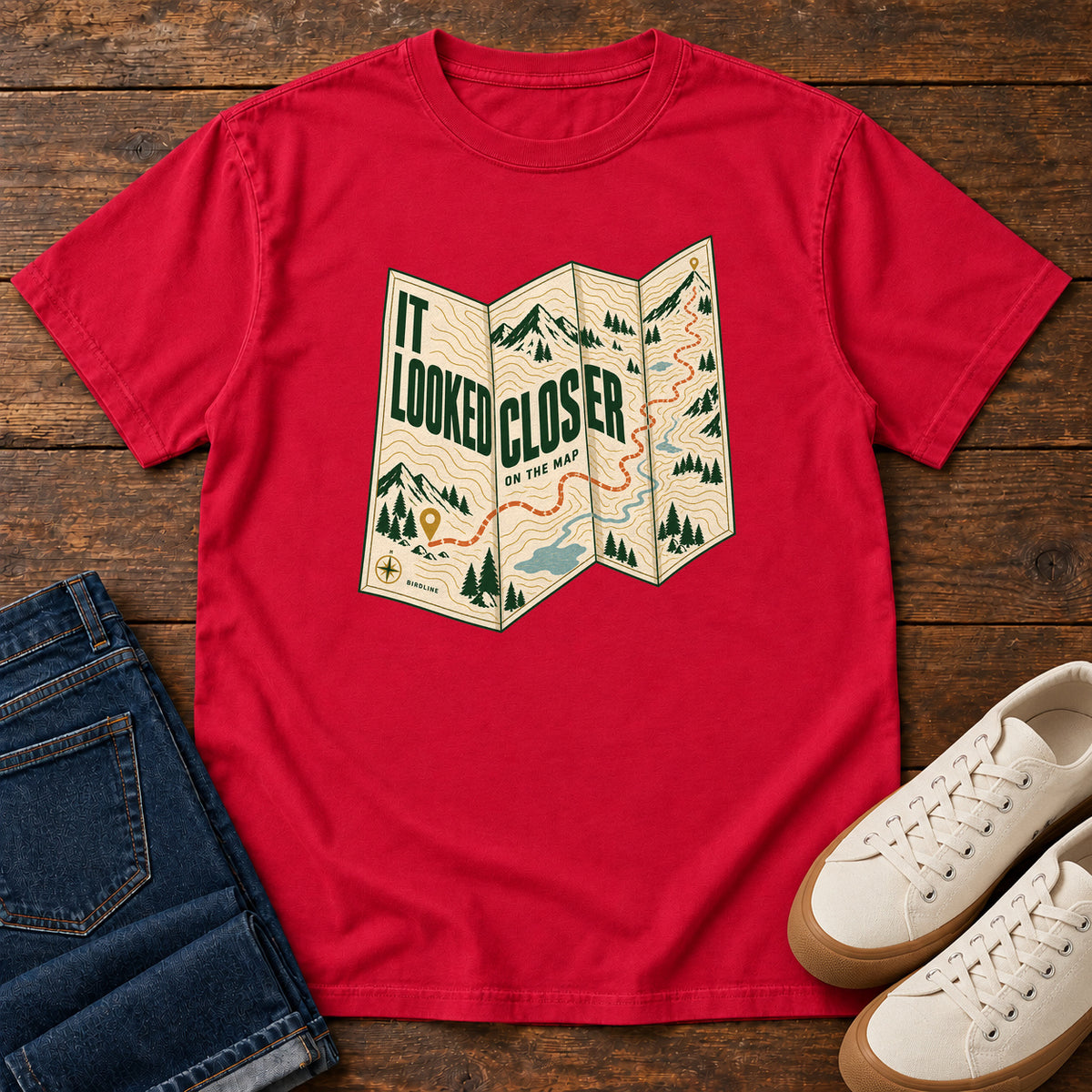 Red It Looked Closer on the Map T-Shirt with a folded cream map graphic, green lettering and an orange route, front flat lay on wood.