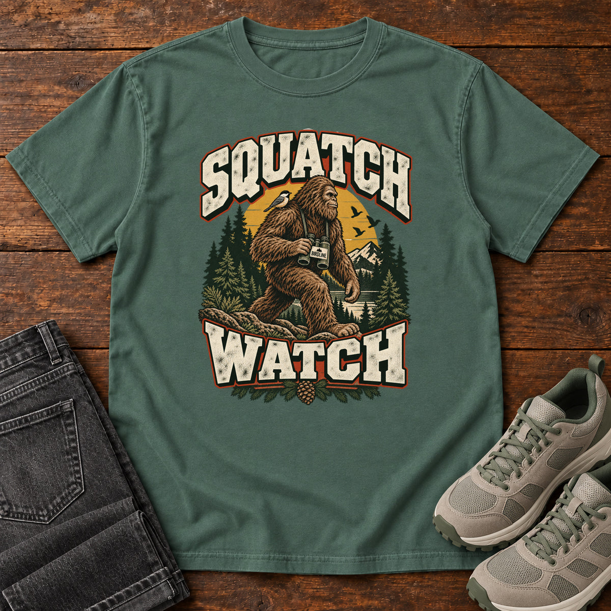 Squatch Watch T-Shirt in Blue Spruce, front laid flat on wood, featuring a walking Sasquatch holding binoculars among pine trees, framed by large cream lettering.