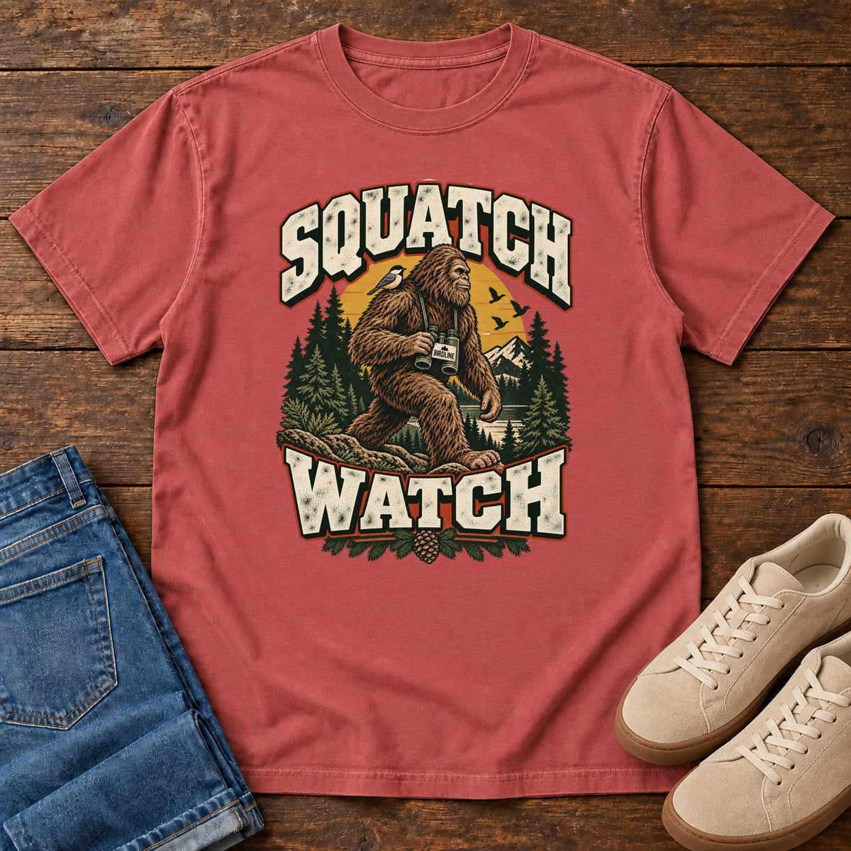 Squatch Watch T-Shirt in Crimson, front laid flat on wood, featuring a walking Sasquatch holding binoculars among pine trees, framed by large cream lettering.
