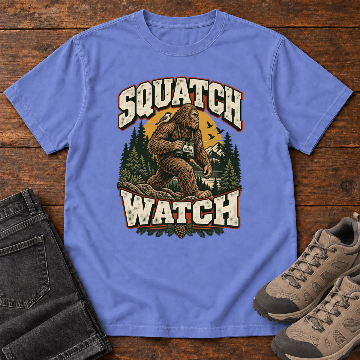 Squatch Watch T-Shirt in Flo Blue, front laid flat on wood, featuring a walking Sasquatch holding binoculars among pine trees, framed by large cream lettering.