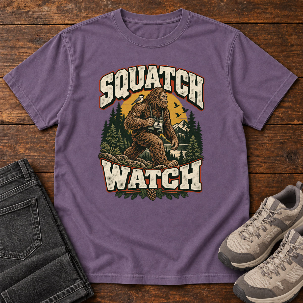 Squatch Watch T-Shirt in Grape, front laid flat on wood, featuring a walking Sasquatch holding binoculars among pine trees, framed by large cream lettering.